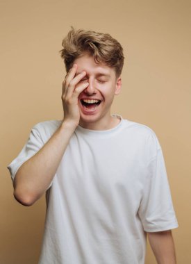 Beyaz tişört giyen genç bir adam yüzünün bir kısmını tek eliyle örterken neşeyle gülüyor. Onun şakacı ifadesi tarafsız bej arkaplana sıcaklık getiriyor.