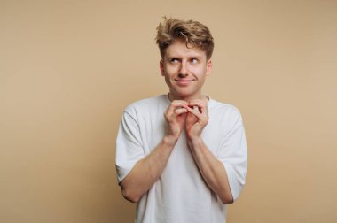 Beyaz gömlekli genç bir adam şakacı bir gülümseme ile duruyor, hafifçe yaramaz görünüyor. Elleri birbirine kenetlenmiş ve açık kahverengi bir arka planı var. Eğlenceli bir atmosfer yaratıyor..