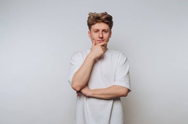 Genç bir adam eli çenesinde, düşünceli bir şekilde ayakta duruyor. Sıradan beyaz bir tişört giyiyor ve spor yapıyor. Rahat bir saç stili, düz bir zemine dayalı..