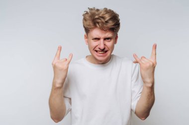 Genç bir adam düz bir zemine karşı duruyor, enerjik bir ifade sergiliyor ve iki eliyle bir kaya işareti yapıyor. Onun tarzı gündelik ve modern..