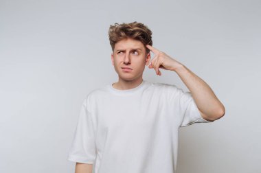 Genç bir adam açık gri bir duvara karşı duruyor, dalgın dalgın bakıyor. Bir eliyle alnına dokunuyor, derin düşüncelere dalmış, beyaz tişört giymiş..