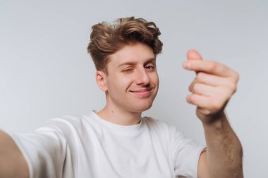 Genç bir adam rahat bir poz veriyor, neşeli bir gülümsemeyle kameraya göz kırpıyor ve eliyle bir kalp şekli oluşturuyor. Arkaplan basit ve hafif.