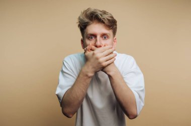 Genç bir adam arka planda durur, iki eliyle ağzını kapatarak şaşkınlık ya da şok olduğunu ifade eder. Geniş gözleri güçlü duyguları açığa çıkarıyor..