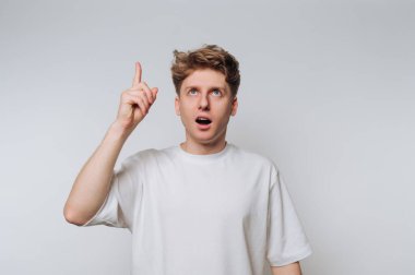 Açık renk saçlı genç bir adam parlak bir stüdyoda duruyor, şaşırmış bir yüz ifadesiyle yukarıya bakıyor. Jesti bir ilham ya da keşif anı olduğunu gösteriyor..