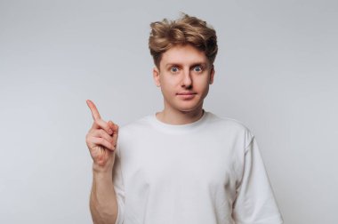 Genç bir adam, işaret parmağını kaldırırken şaşırmış bir şekilde sade bir arkaplanda duruyor. İfadesi bir şeyin farkına vardığını ya da harika bir fikir olduğunu gösteriyor..
