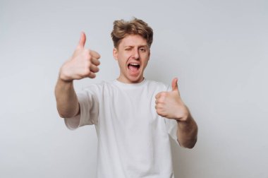 Genç bir adam sade bir arka planın önünde duruyor, coşkusunu büyük bir gülümseme ve iki başparmağıyla gösteriyor. Neşeli bir şekilde göz kırpar, bir mutluluk anı yakalar..