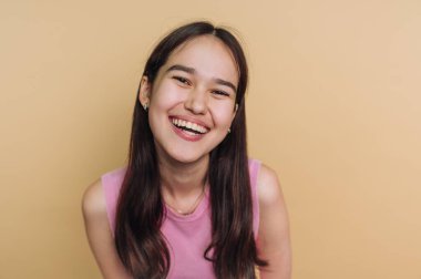 Siyah, uzun saçlı mutlu bir genç kız hafifçe öne doğru eğilerek geniş bir şekilde gülümsüyor. Pembe kolsuz bir bluz giyiyor, neşeli ve dostane bir atmosfer yaratıyor..