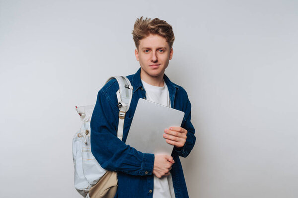 A young man stands confidently with a laptop in hand and a backpack slung over one shoulder. He poses against a plain background, showcasing a casual style.