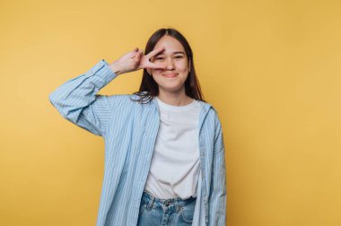 Sarı arka planda barış işareti yapan genç bir kadın gülümsüyor ve sıradan bir kıyafet giyiyor.