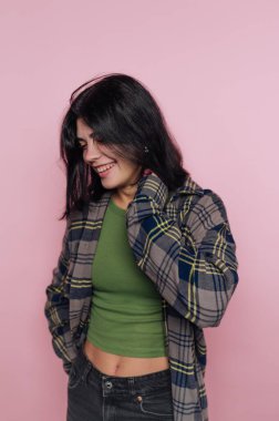 Ekose gömlekli, yeşil bluzlu, pembe arka planda gülümseyen bir kadın..