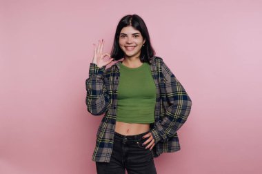 Gündelik giyinmiş genç bir kadın pembe bir arka plana karşı iyi bir jest yapıyor..