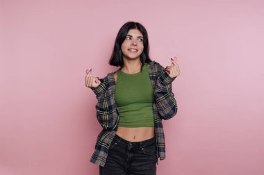 Gündelik kıyafetli genç bir kadın pembe arka planda parmak kalp hareketleri yapıyor..