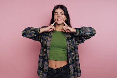 Gülümseyen ve şakacı bir jest yapan genç bir kadın, parmakları yanaklarında, yeşil bir bluz ve ekose bir gömlek giyiyor, pembe bir arka plana karşı