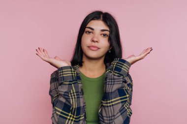 Kafası karışmış, ekose gömlekli, yeşil bluzlu, pembe arka planda duran genç bir kadın..