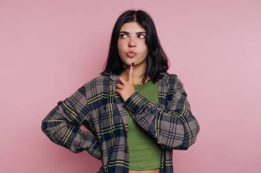 Ekose gömlekli genç bir kadın pembe arka planda parmakları çenesinde düşünüyor..