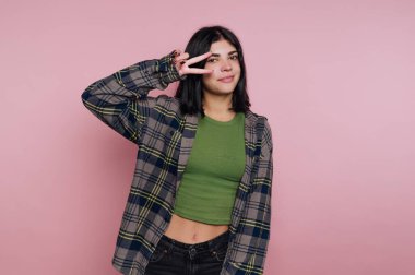 Gündelik giyinmiş genç bir kadın pembe arka planda barış işareti yapıyor..