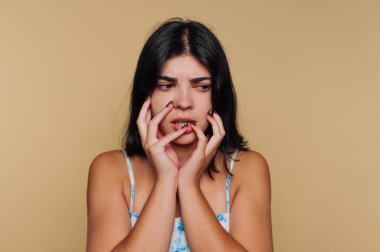 Koyu renk saçlı, çiçekli elbisesi bej arka planda olan endişeli bir kadın..