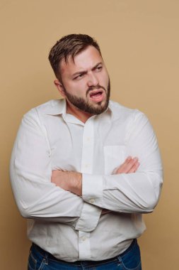 Sakallı ve bıyıklı bir adam, beyaz bir gömlek giymiş, kolları bej bir arka planda çapraz duran, kafası karışmış ya da kafası karışmış görünen..