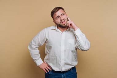 Beyaz gömlekli ve kot pantolonlu bir adam elleri çenesinde bej bir arka planda düşünüyor.