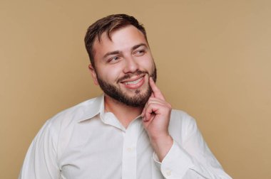 Beyaz tişörtlü sakallı bir adam, gülümsüyor ve bej bir arka plana karşı düşünüyor.