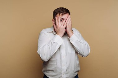 Beyaz gömlekli bir adam elleriyle yüzünü kapatıyor, şaşırmış görünüyor..