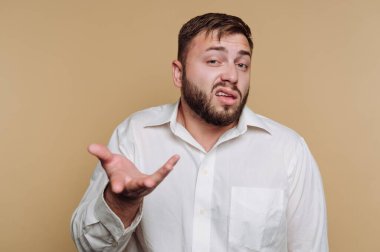 Beyaz gömlekli bir adam bej arka planda karışık bir ifadeyle omuz silkiyor..