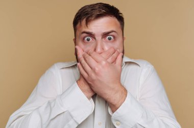 Şaşırmış bir adam bej arka planda iki eliyle ağzını kapatır..