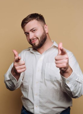 Beyaz gömlekli bir adam bej arka planda parmak tabancasıyla şakacı poz veriyor.