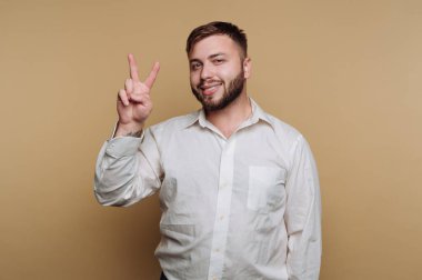 Beyaz gömlekli gülümseyen adam bej arka planda barış işareti yapıyor..