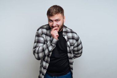 Ekose gömlekli genç bir adam düz bir arka planda duruyor, oyuncu ve biraz utangaç görünüyor, bir eli ağzının yanında.