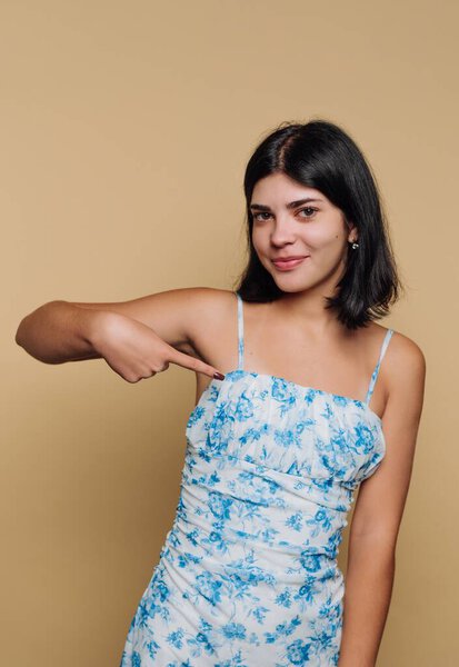 Женщина в цветочном платье указывает на себя на бежевом фоне.