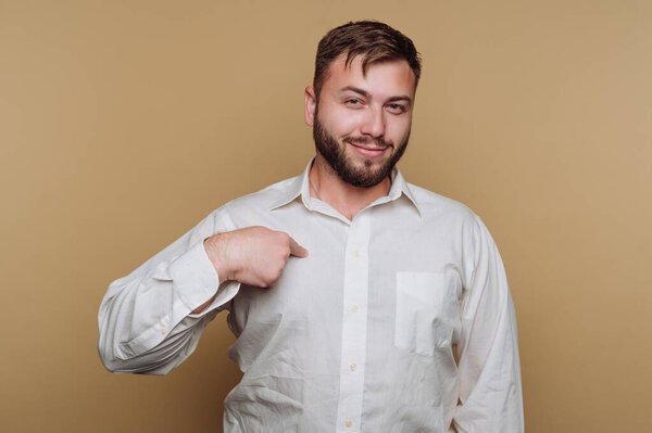 Человек в белой рубашке указывает на себя на бежевом фоне.