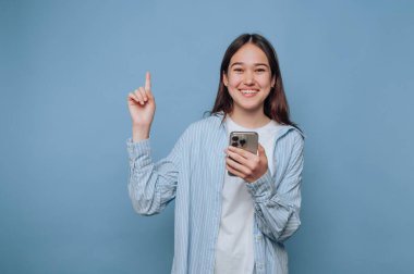 Elinde akıllı bir telefonla gülümseyen genç bir kadın mavi bir arka plana doğru bakıyor.