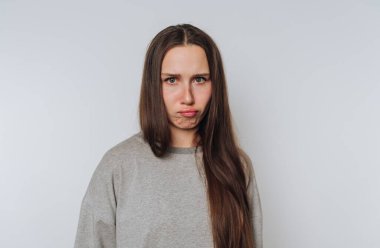 Uzun saçlı genç bir kadın, gri bir arka planda üzgün bir surat yapıyor..