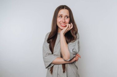 Endişeli bir kadın tırnaklarını yiyor, gri bir gömlek giyiyor, arka planda..