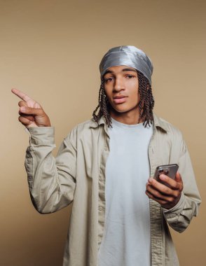 Saçları örgülü, durag takan, elinde akıllı bir telefon tutan ve bej arka planı olan genç bir adam..