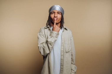 Düşünceli bir yüzü olan genç bir adam, uzun ve sıradan kıyafetler giyiyor, düz bir bej arka planı var..