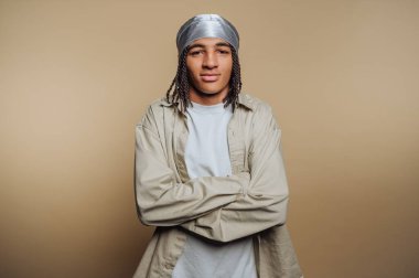 Örgülü saçlı, gümüş rengi başörtülü, beyaz tişörtlü, kolları bej arka planda çapraz duran genç bir adam..