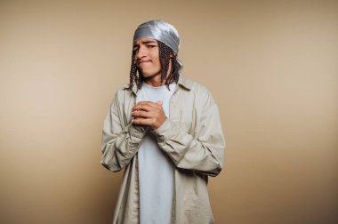 Saçları örgülü, günlük giysiler giymiş bej bir arka plana karşı duran düşünceli bir genç..