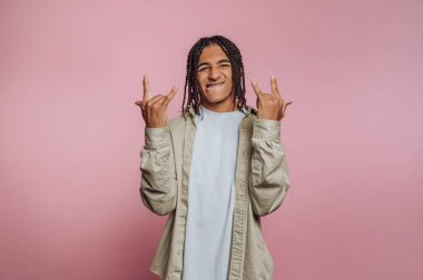 Saçları örgülü genç bir adam pembe arka planda kaya el işaretleri yapıyor..