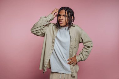 Saçları örgülü genç adam pembe arka planda kafası karışmış görünüyor..