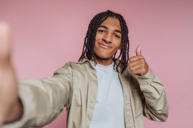 Saçları örgülü genç adam pembe arka planda başparmak işareti yapıyor..