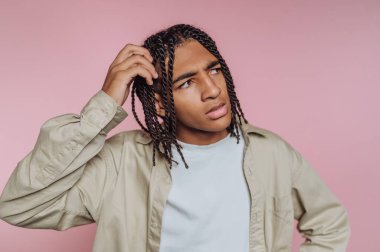 Saçları örgülü genç adam pembe arka planda kafası karışmış görünüyor..