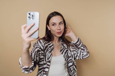 Bej arka planda akıllı telefonuyla selfie çeken bir kadın..