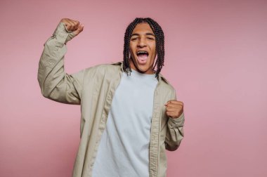 Genç adam yumruklarıyla kutluyor, gündelik kıyafetler giyiyor, pembe arka plan.