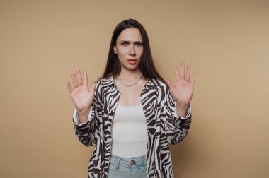 Uzun saçlı, zebra desenli tişörtlü bir kadın bej arka planda el kaldırıyor..