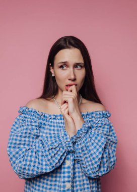 Mavi kareli bluzlu kadın parmağını ısırıyor, pembe arka plan.