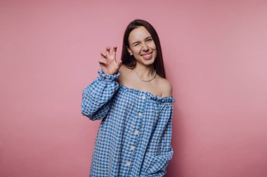 Mavi kareli elbiseli kadın pembe arka planda şakacı poz veriyor..