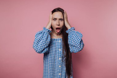 Pembe arka planda, mavi kareli elbiseli, uzun saçlı, şaşırmış bir kadın..