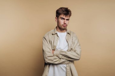 Çapraz kollu genç bir adam ve bej bir arka plana karşı ciddi bir ifade.